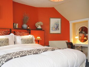 A bedroom with a bed, pillows, a nightstand, a lamp, a radiator, a mirror, and a shelf with decorative items. - Sycamore Apartment (Lifton)