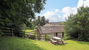 A rustic stone house with a thatched roof, a wooden fence, and a picnic table in a lush green garden. - Abbots Reading Mews 2 (Ulverston)