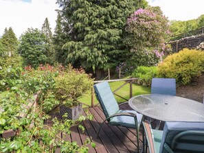 A wooden deck with a glass table and two chairs, surrounded by lush greenery and flowering plants. - The Farmhouse (Coniston)