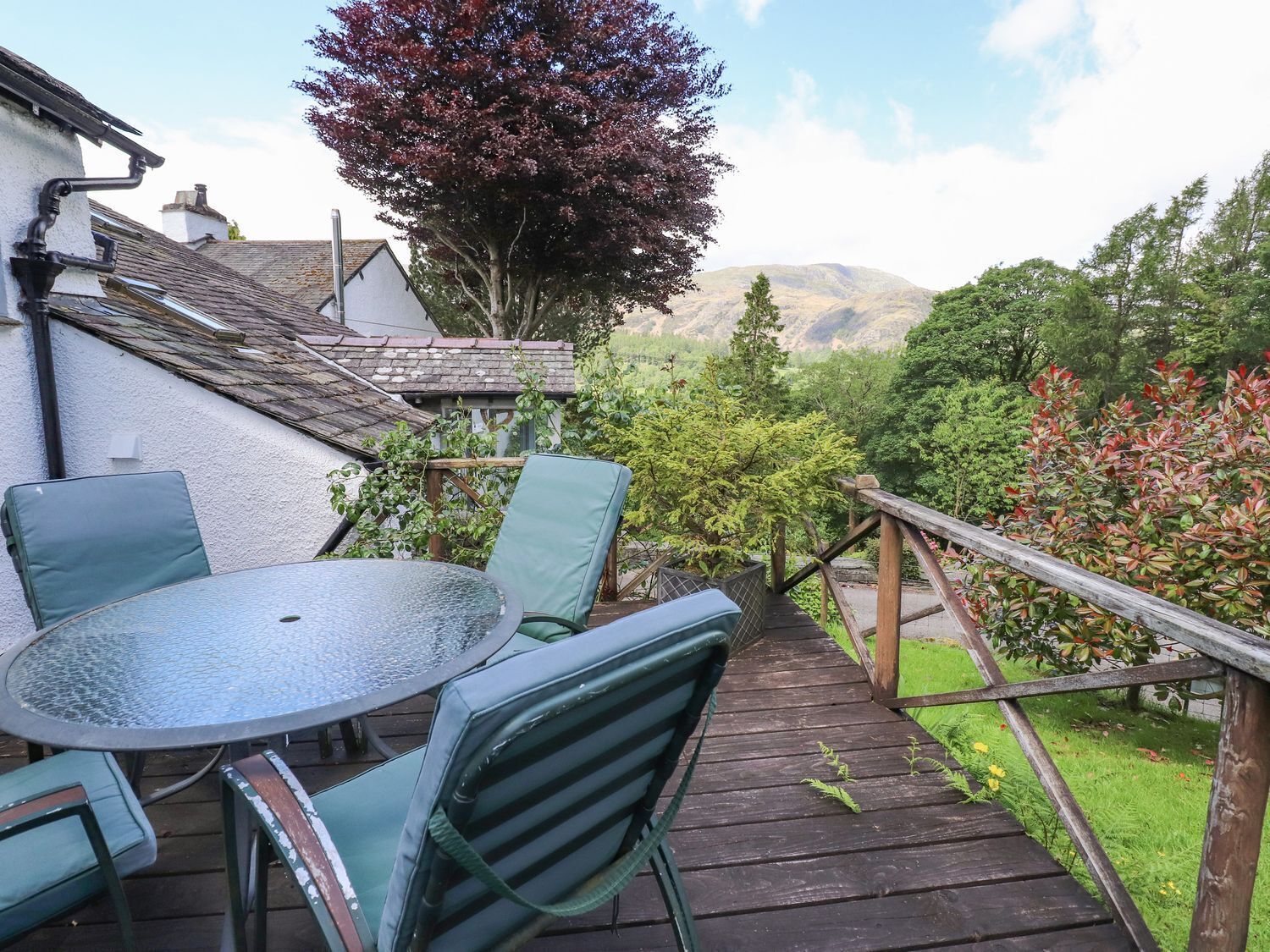 A wooden deck with a glass table and chairs, surrounded by lush greenery and a view of hills.