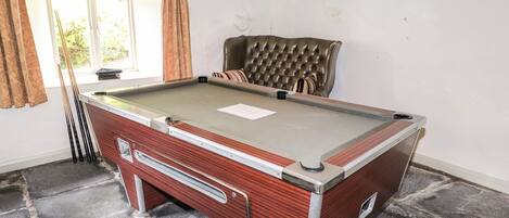 A pool table with a wooden frame, a cushioned chair, and a window with curtains.