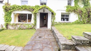 A white house with a stone pathway leading to the entrance, surrounded by greenery and a stone wall. - The Farmhouse (Coniston)