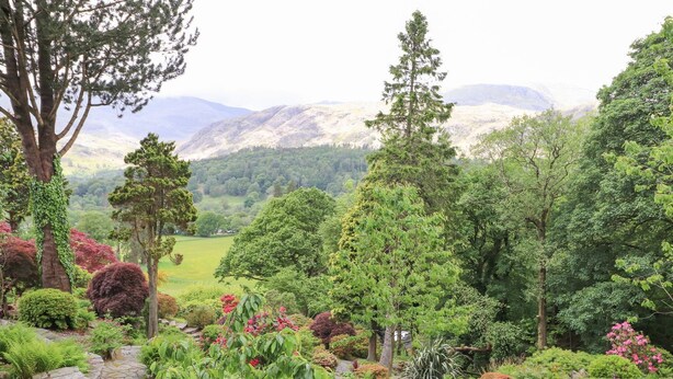 A lush garden with various trees and shrubs, a stone pathway, and a view of distant mountains. - The Farmhouse (Coniston)