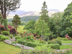 A lush garden with various trees and shrubs, a stone pathway, and a view of distant mountains. - The Farmhouse (Coniston)