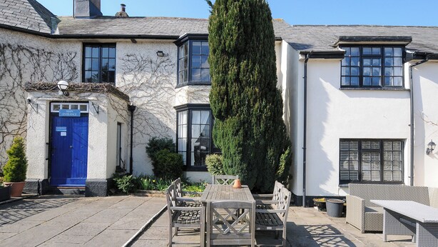 A courtyard with a blue door labeled "Main Entrance," a wooden table with chairs, and a tall coniferous tree. - Alway (Lifton)