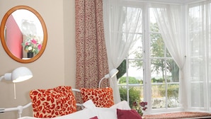 A bedroom with a bed, a bedside lamp, a window with sheer curtains, and a view of greenery outside. - Alway (Lifton)