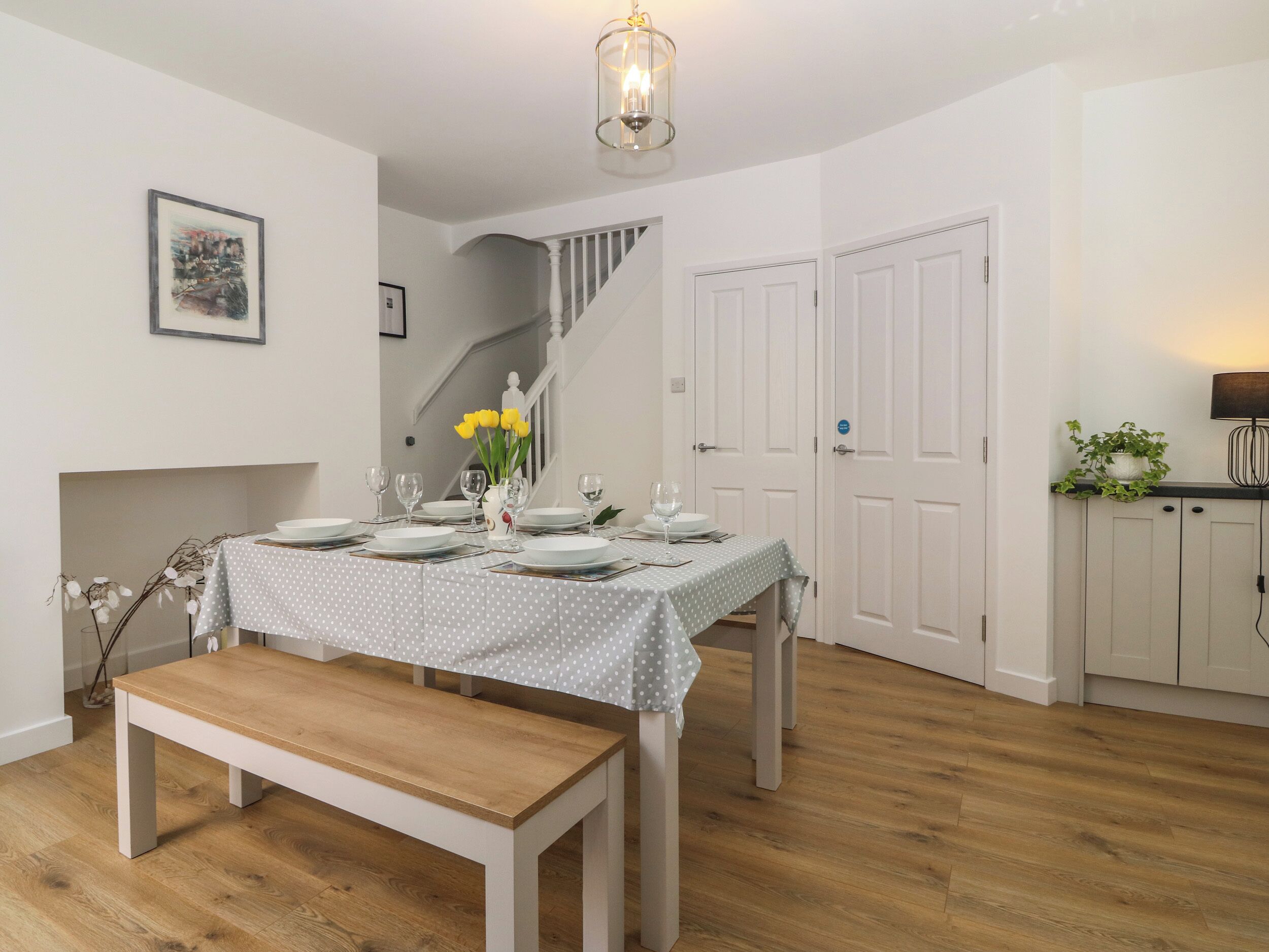 Un comedor con un banco de madera, una mesa dispuesta con platos, vasos y un florero. Se ve una escalera al fondo, que conduce a otra habitación.
