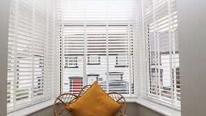 A corner room with white blinds, a wicker chair, and a view of buildings outside. - Elsinore (Conwy)