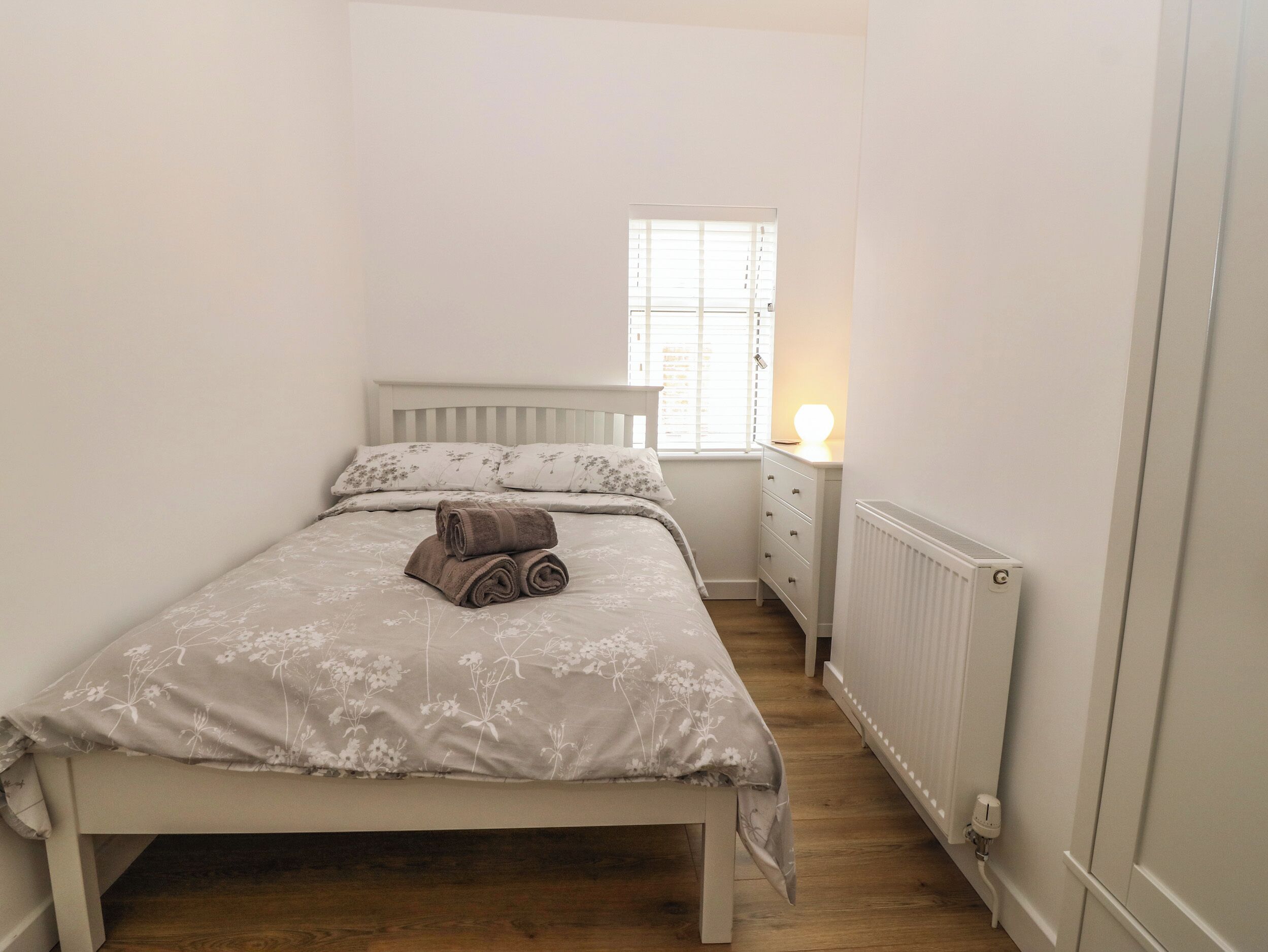 A small, neatly arranged bedroom with a single bed, a nightstand, a dresser, and a window with blinds.