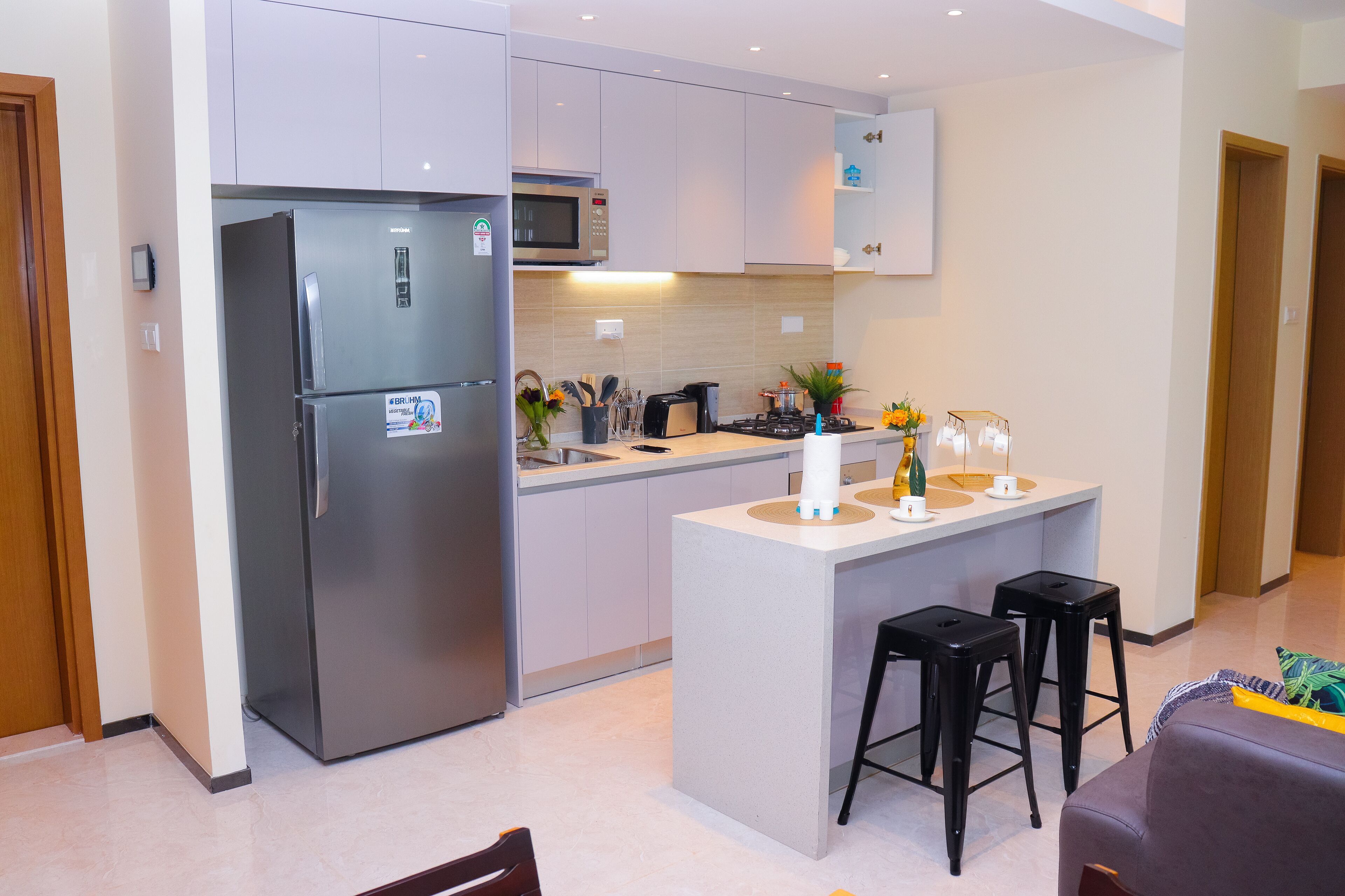 A modern kitchen with a built-in refrigerator, microwave, and stove. There are two black bar stools and a small island with a sink.