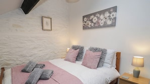 A bedroom with a stone wall, a bed with pink and grey pillows, a bedside table with a lamp, and a painting on the wall.