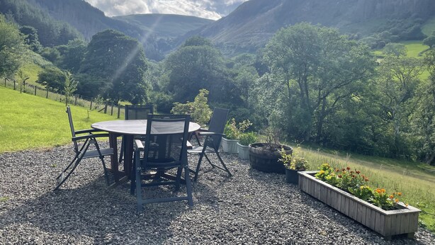 An outdoor seating area with a table and chairs, surrounded by gravel and potted plants, overlooking a valley with mountains and greenery.