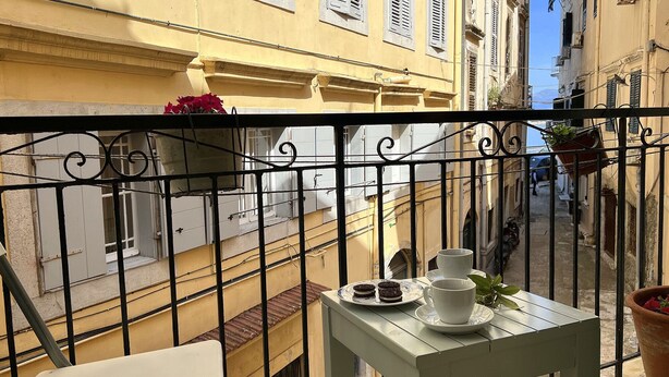 A balcony with a table set for tea, overlooking a narrow street with buildings. - Casa Di Porto (Kerkira)