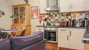 A compact kitchen with white cabinets, a tiled backsplash, and a built-in oven. There's a wooden cabinet with glass doors and a green pendant light. - The Barn (Lifton)