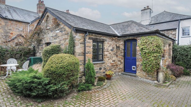 A stone building with a blue door, surrounded by greenery and a cobblestone path. - The Barn (Lifton)