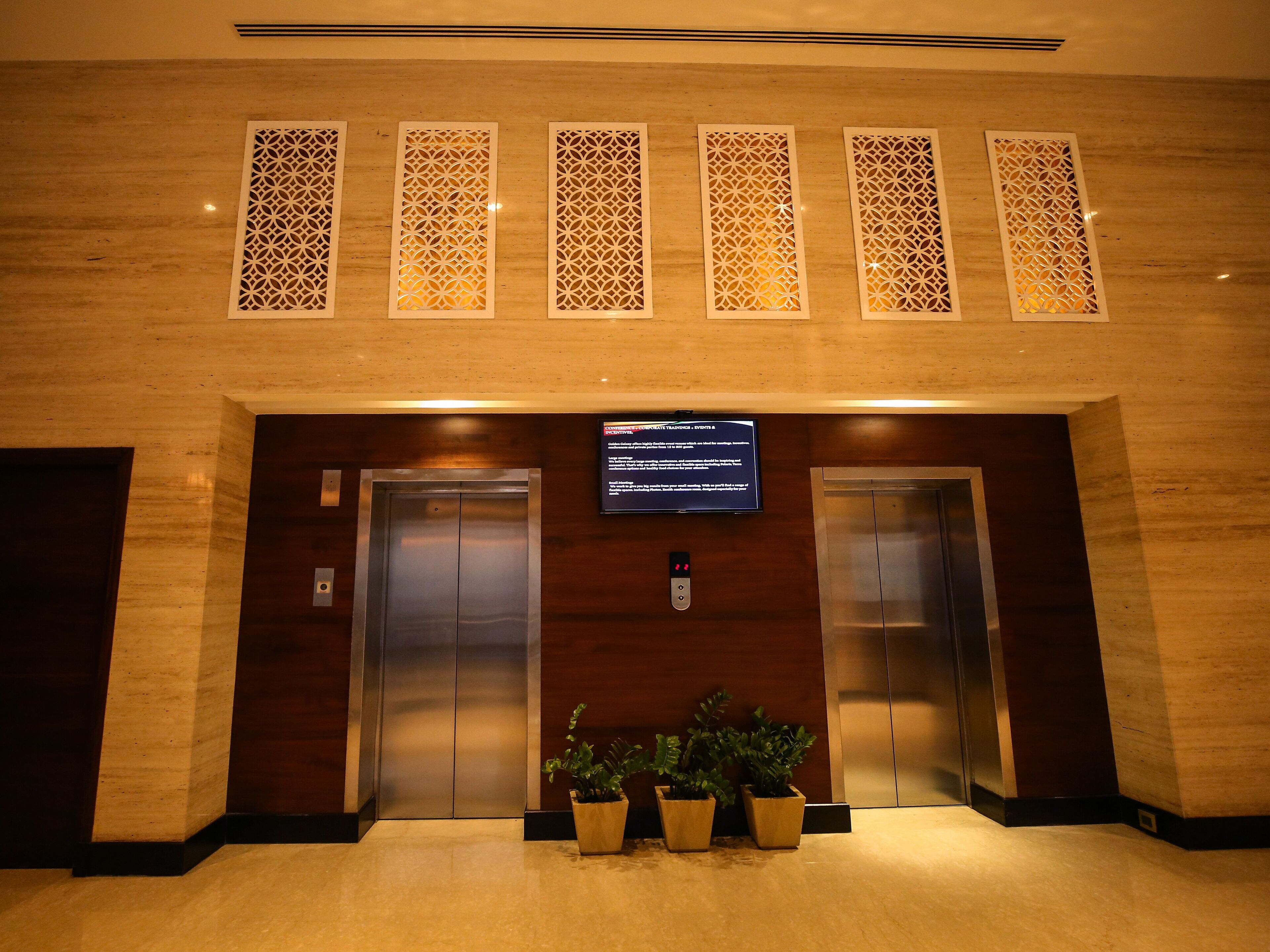 A hotel lobby with two elevators, decorative panels, a digital information board, and potted plants.