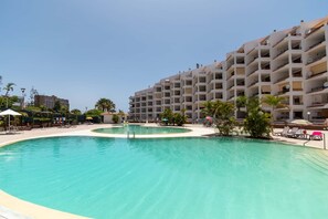 Una amplia piscina de diseño curvo, rodeada de sillones de descanso y sombrillas, junto a un hotel de varios pisos con balcones.
