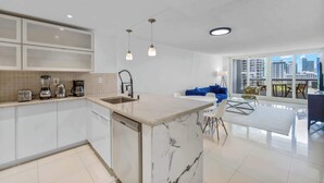 Una cocina moderna con una isla de mármol, armarios blancos y un comedor con sofá azul y vistas a la ciudad.