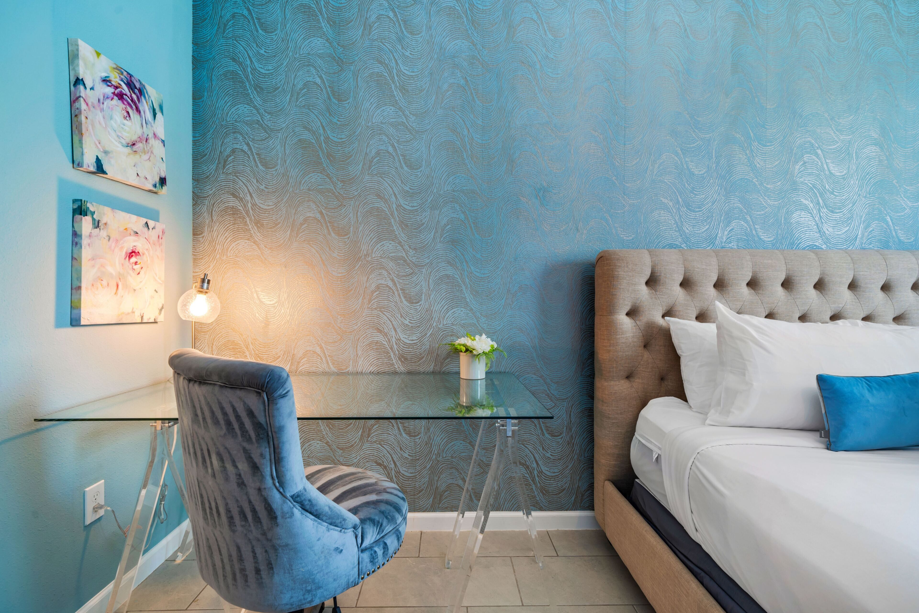 A bedroom with a tufted headboard, a glass table with a potted plant, a blue chair, and artwork on the wall.