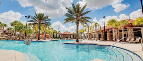 A large outdoor swimming pool with palm trees, lounge chairs, and a covered area with a red roof.