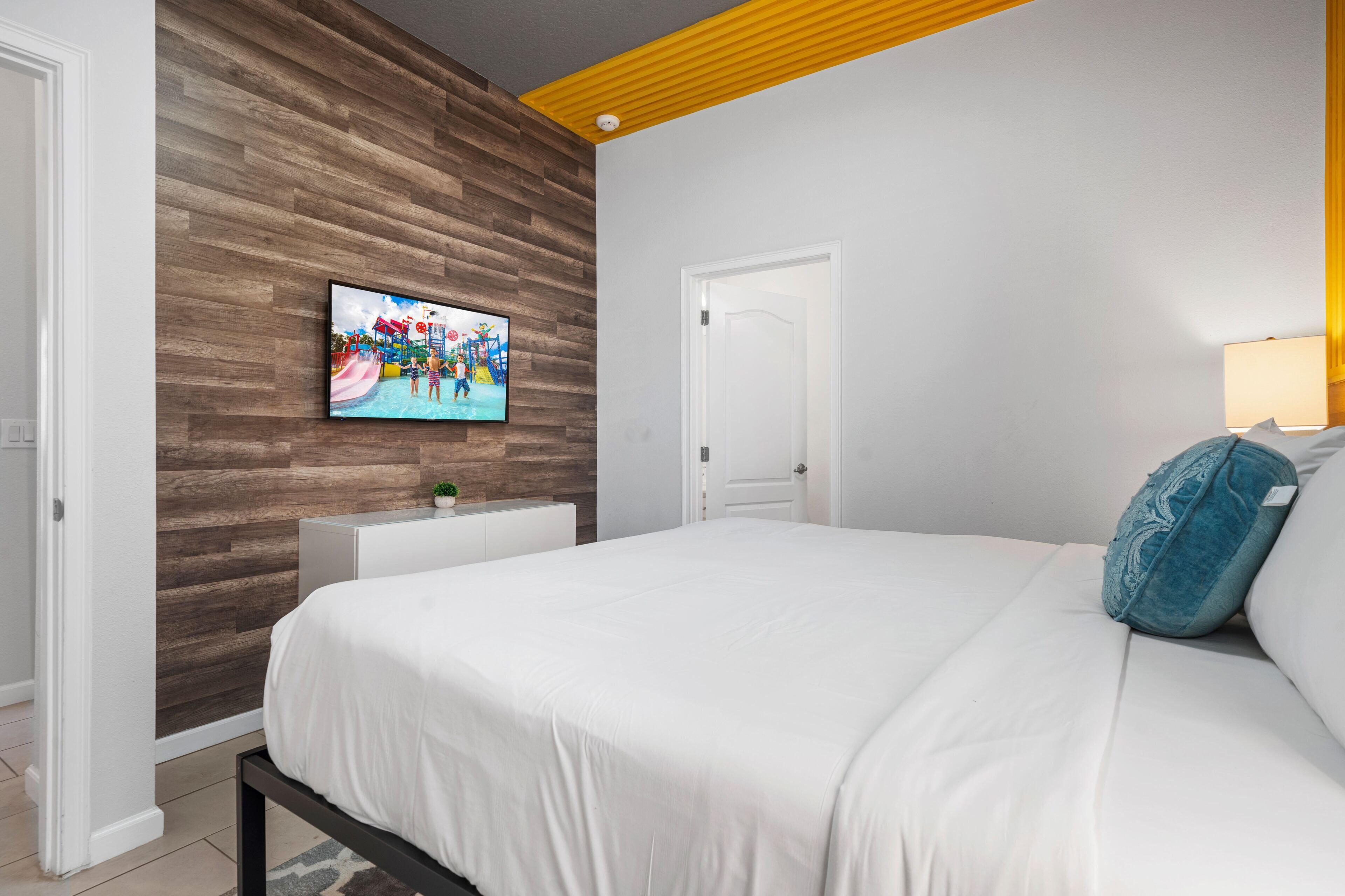 Quarto com cama, televisão fixada numa parede de madeira, mesa de cabeceira branca e uma porta.