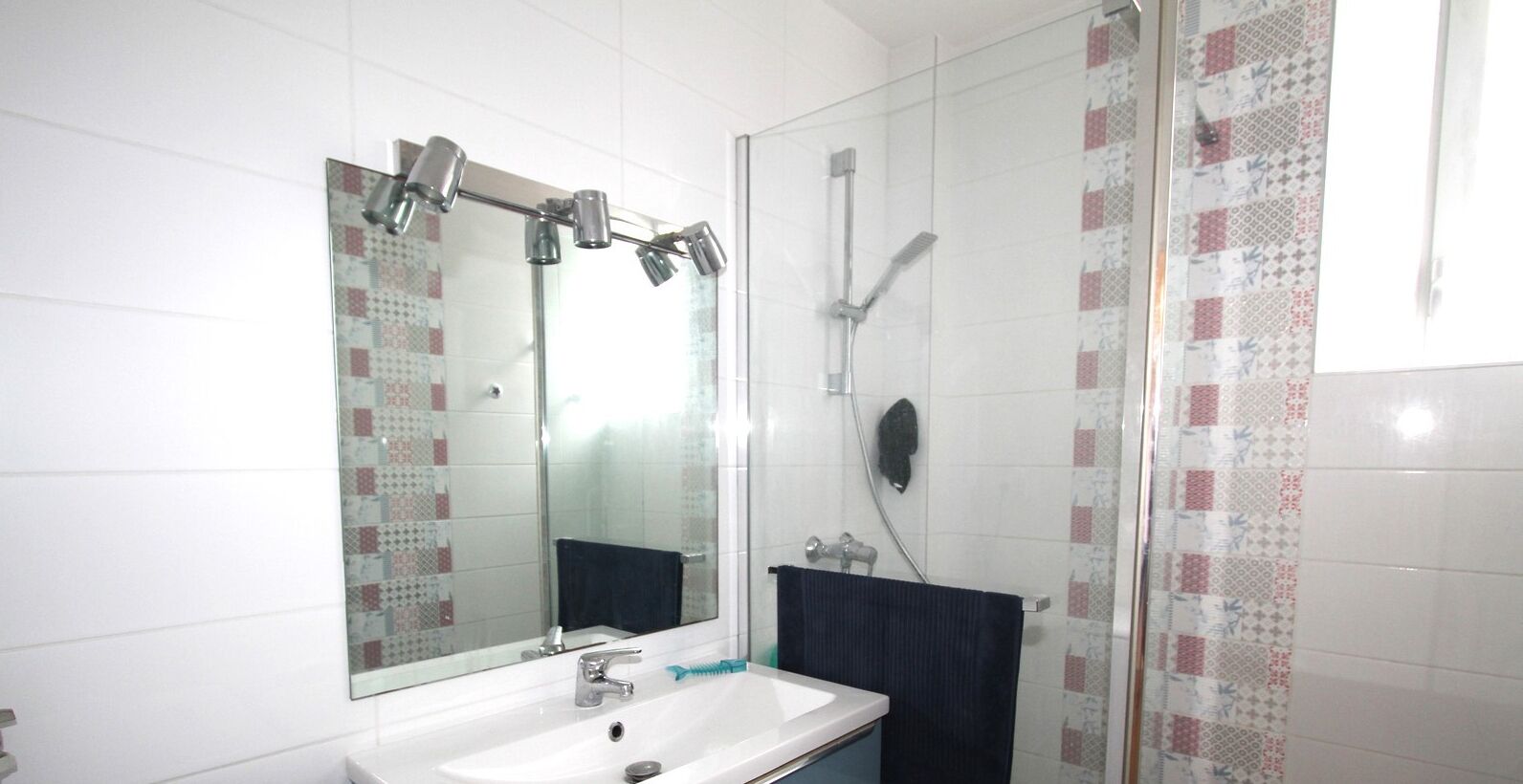 A bathroom with a white sink, a large mirror, a shower with a glass door, and patterned tiles on the walls.