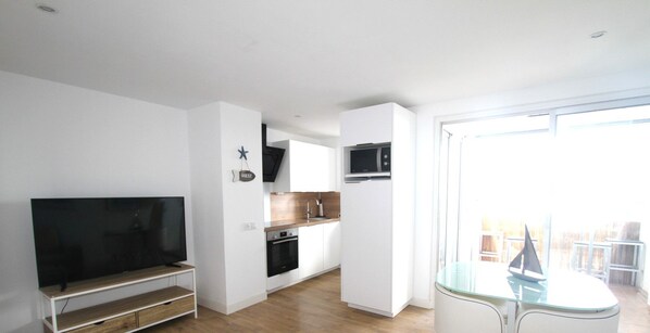 A modern living room with a flat-screen TV, wooden flooring, a glass dining table, and a kitchen area with white cabinets and a microwave.