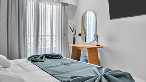 Ein modernes Hotelzimmer mit einem ordentlich bezogenen Bett, einem Holfnachtschränkchen, einem Spiegel und einem großen Fenster mit durchsichtigen Vorhängen.