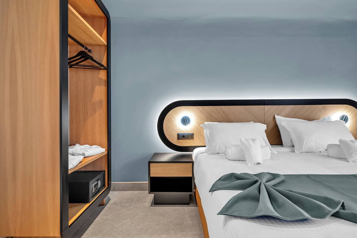 A modern hotel room with a wooden wardrobe, a neatly made bed with white linens, and a bedside table with a lamp.