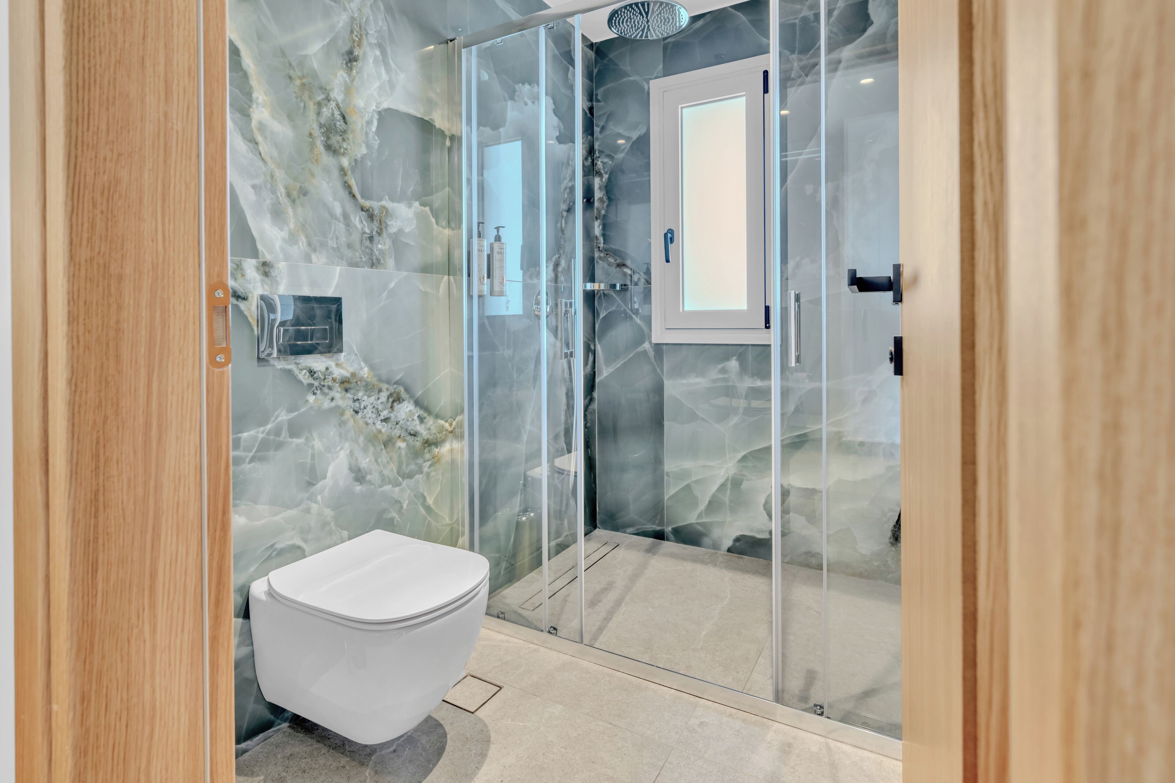Un baño moderno con pared de mármol, ducha con cerramiento de vidrio y un inodoro blanco.