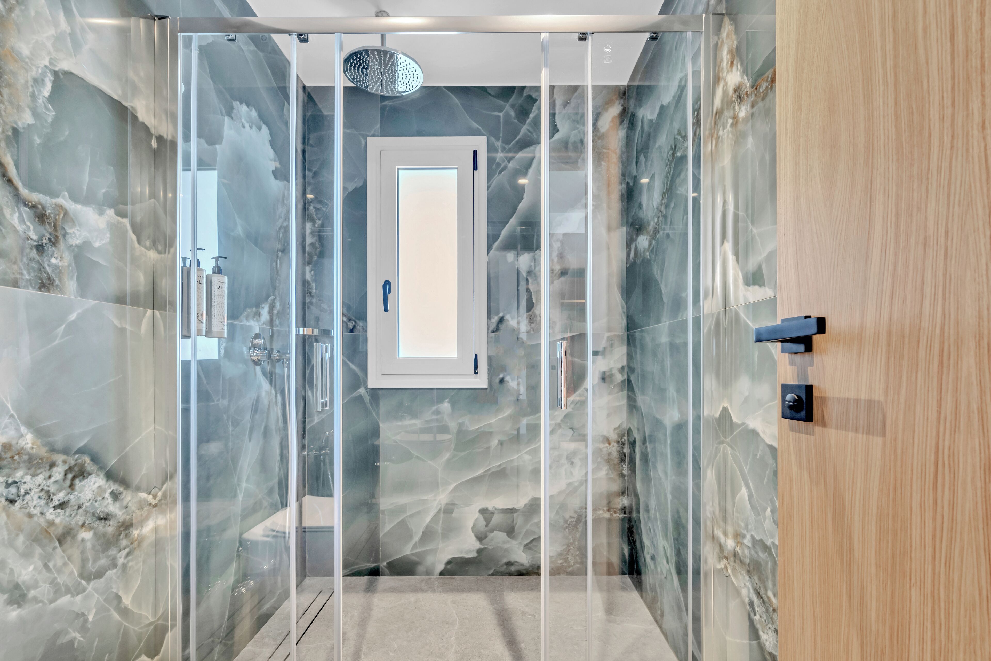 A modern bathroom with a glass-enclosed shower, marble walls, and a wooden door.