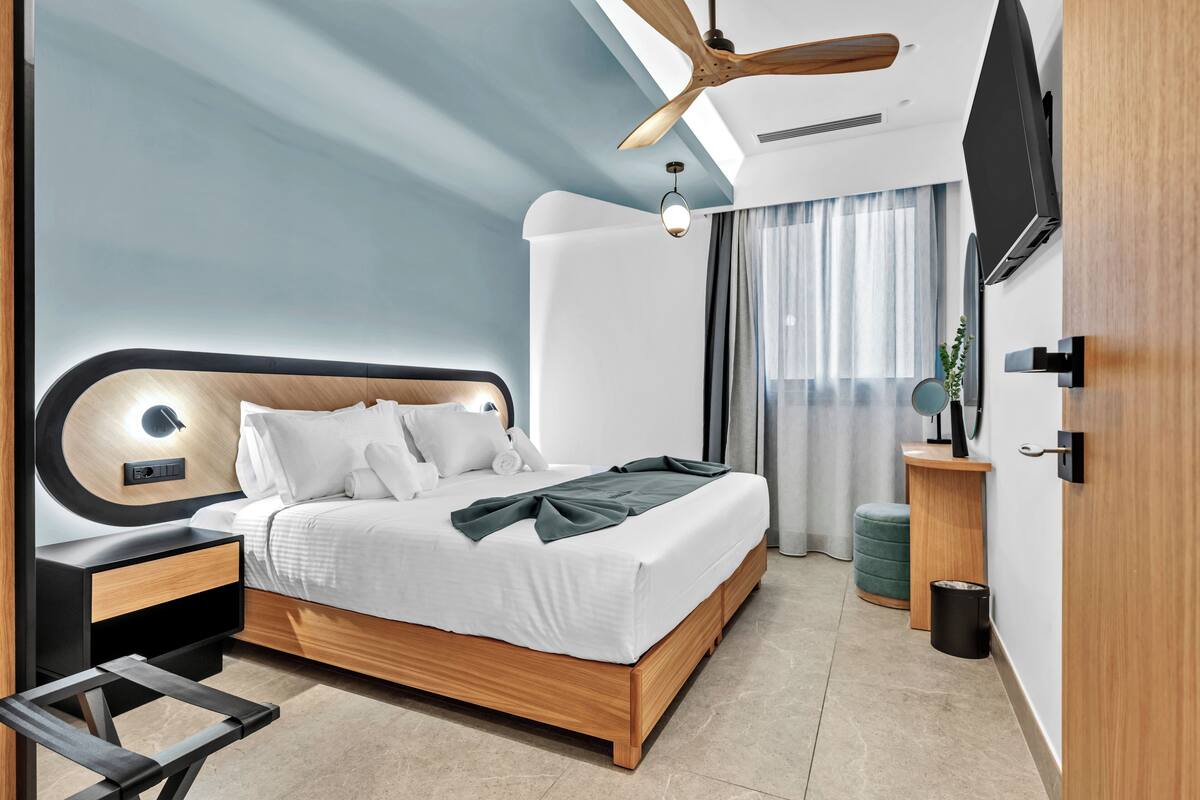 A modern bedroom with a wooden bed, a ceiling fan, a bedside table, a TV mounted on the wall, and a window with curtains.