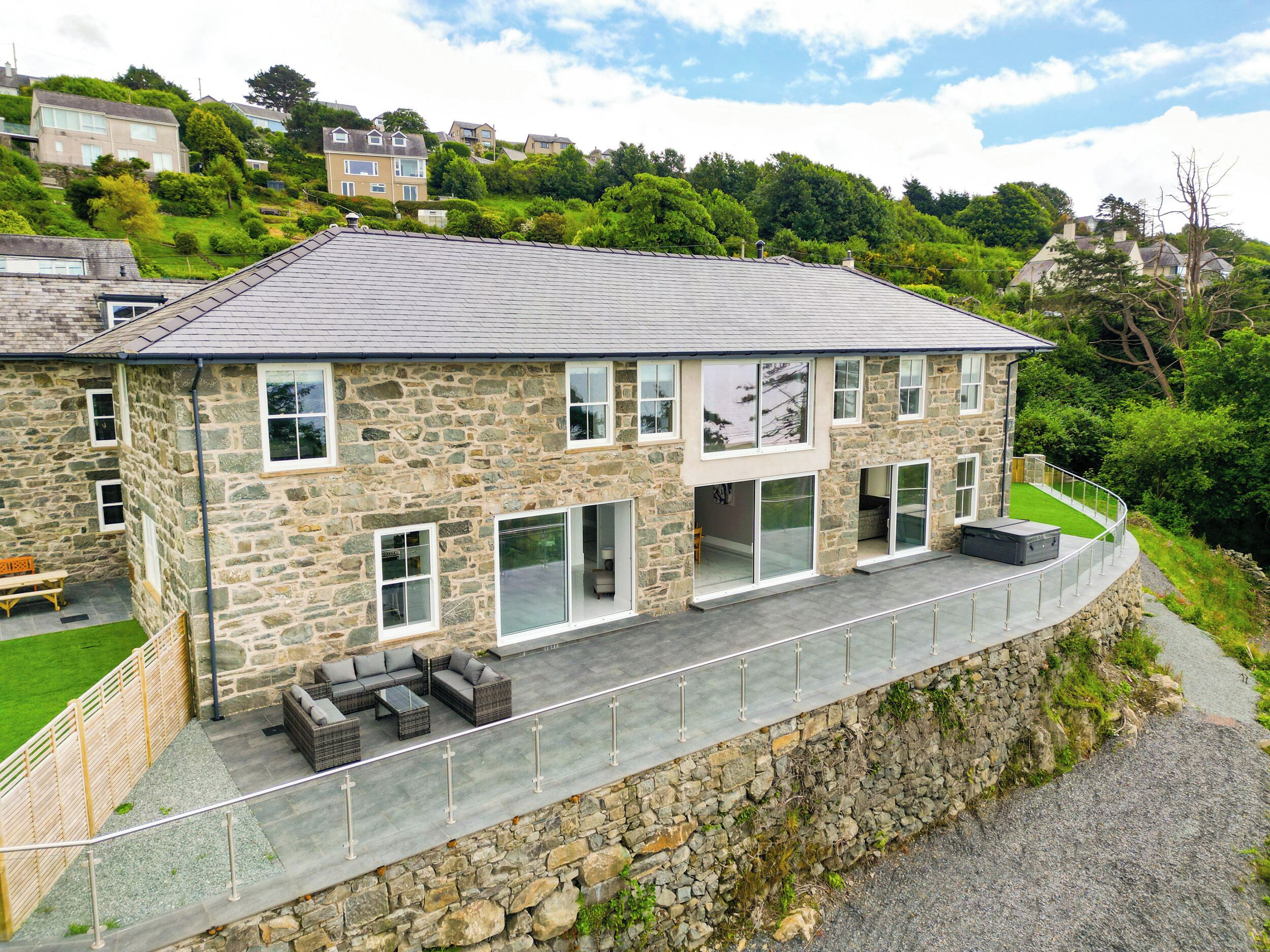 A stone house with a flat roof, large windows, and a balcony with outdoor furniture.