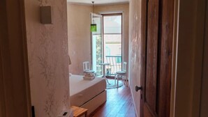 Un pasillo estrecho que conduce a un dormitorio con una cama, una mesita y una silla. Se observa una puerta a la derecha y una ventana a la izquierda.