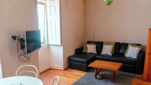 Una sala de estar con un sofá negro, una mesa de centro de madera, una mesa blanca y un televisor montado en la pared.