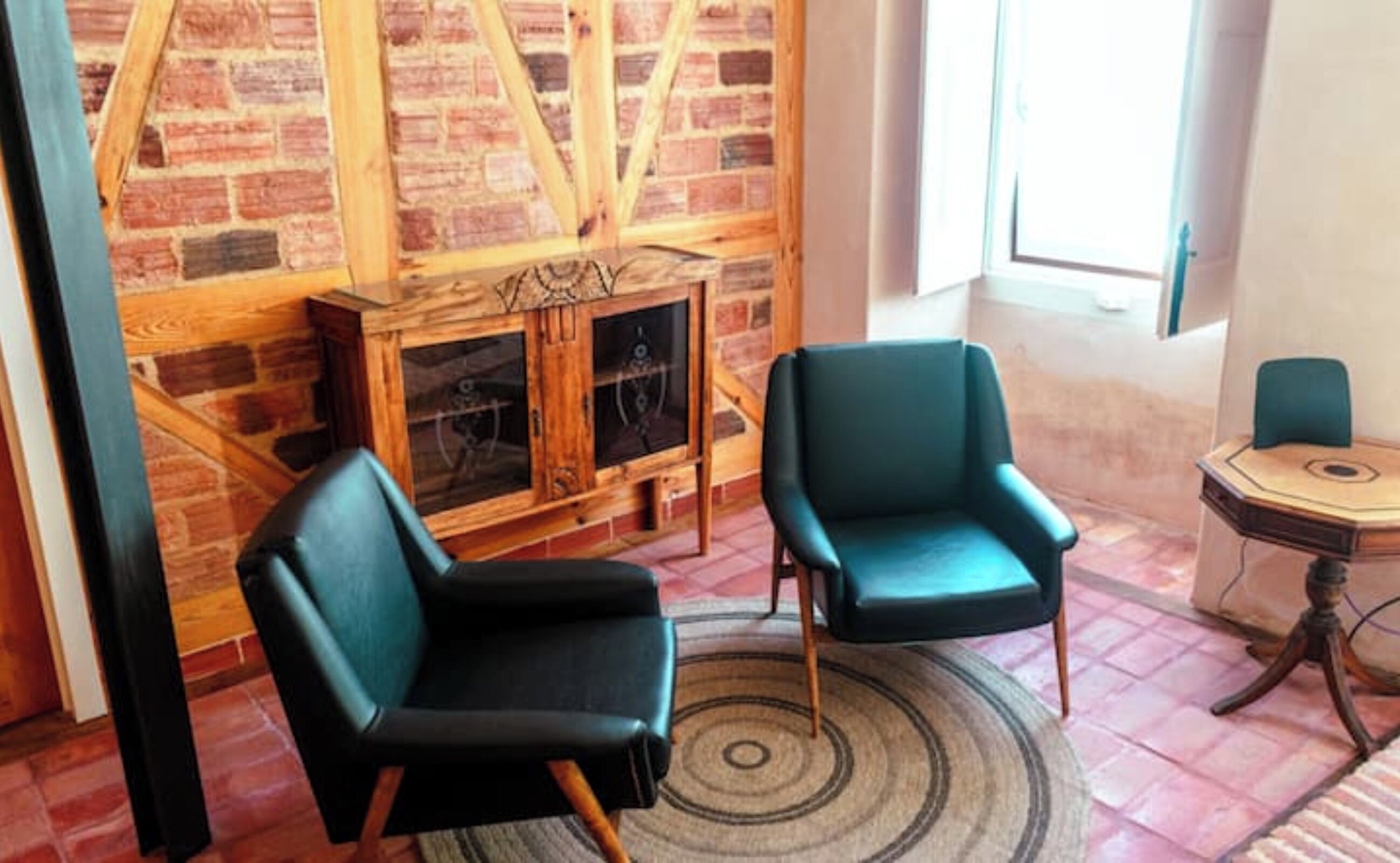 Une pièce avec une cheminée en briques, deux fauteuils de couleur turquoise, un tapis rond et une petite table agrémentée d’un objet décoratif.
