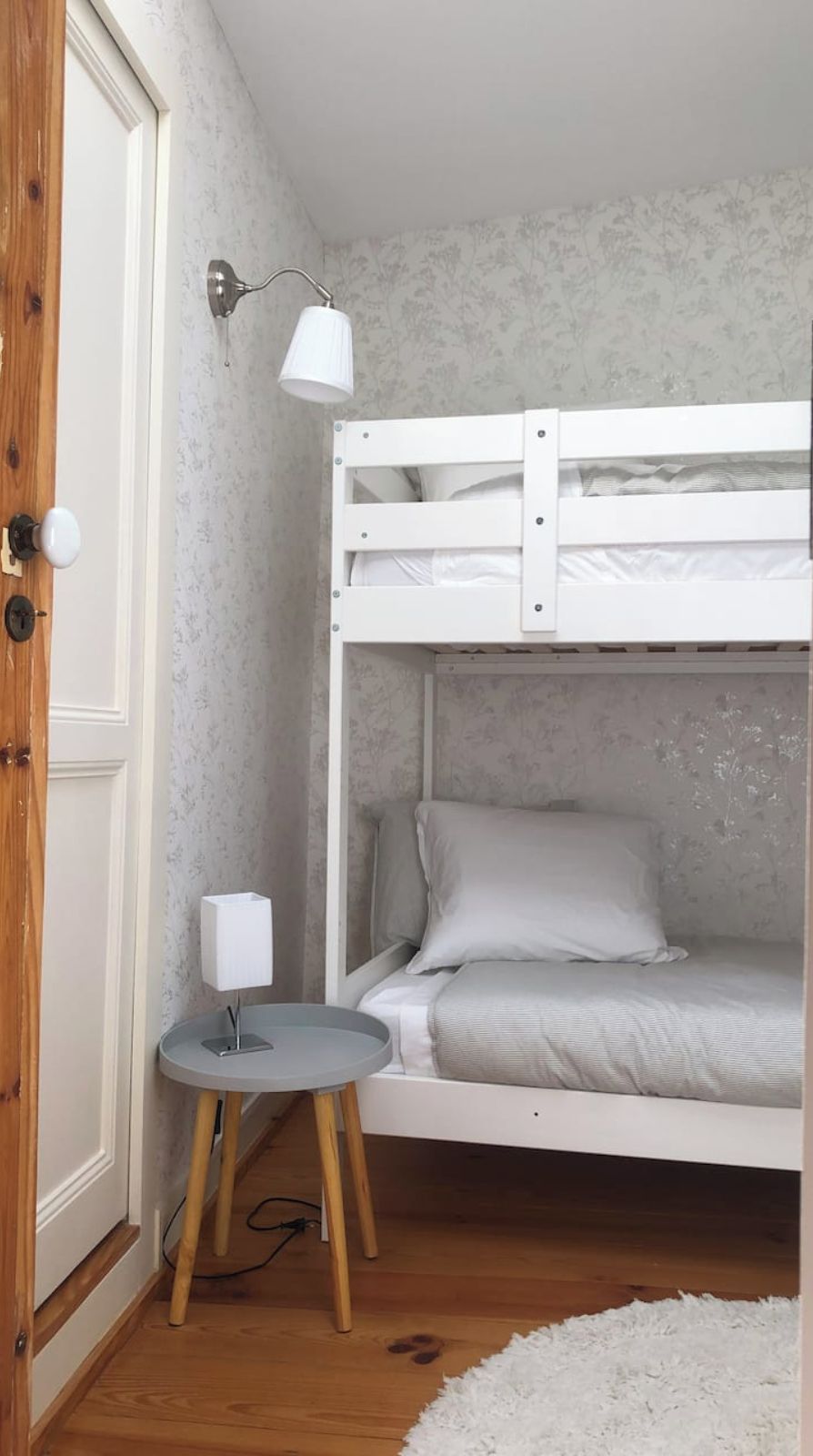 Une chambre avec un lit superposé, une petite table et une lampe, un plancher en bois et une porte blanche.