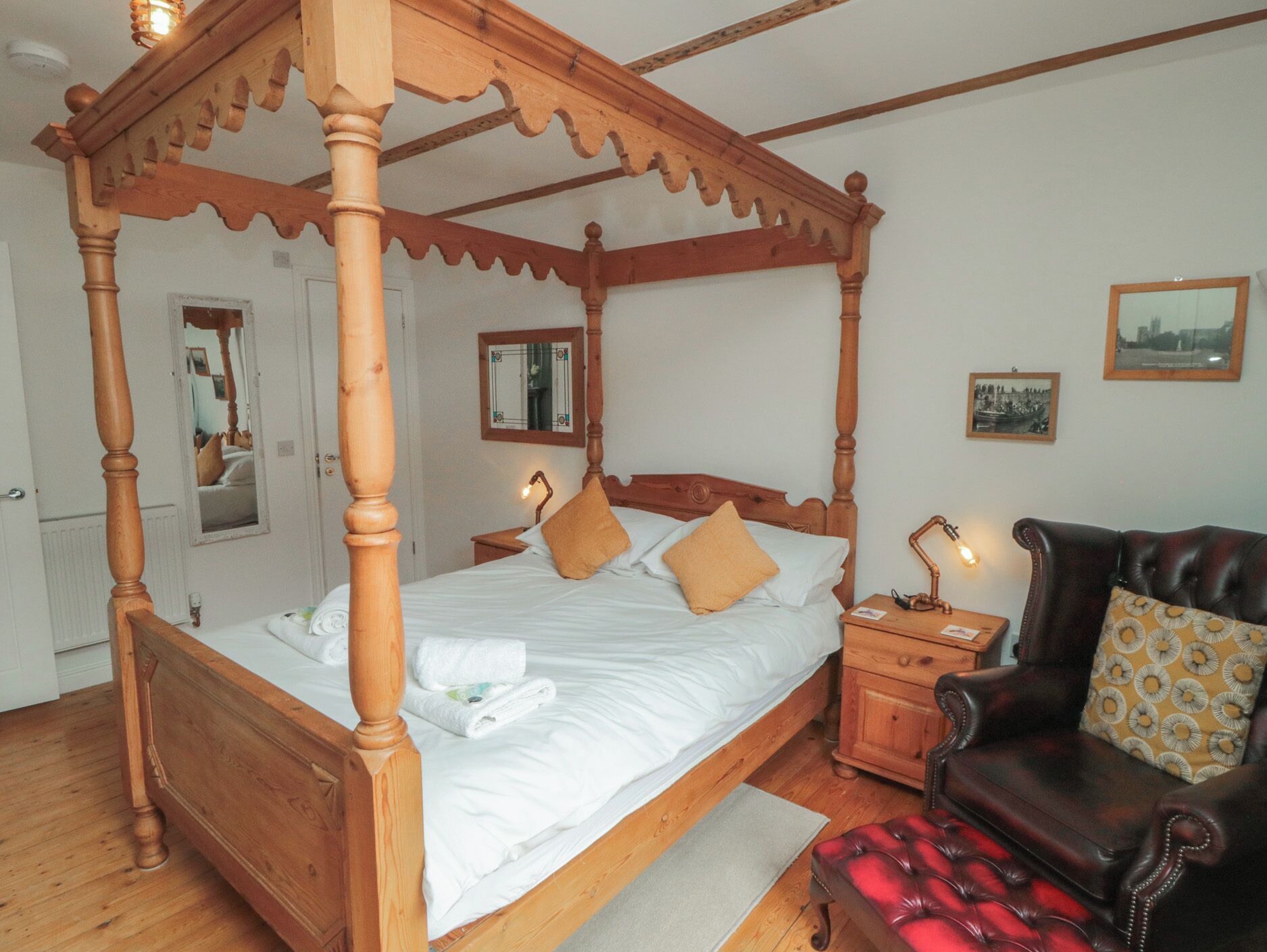 Una cama con dosel de madera, una mesita de noche con lámpara, un sillón de cuero y dos cuadros enmarcados en la pared.