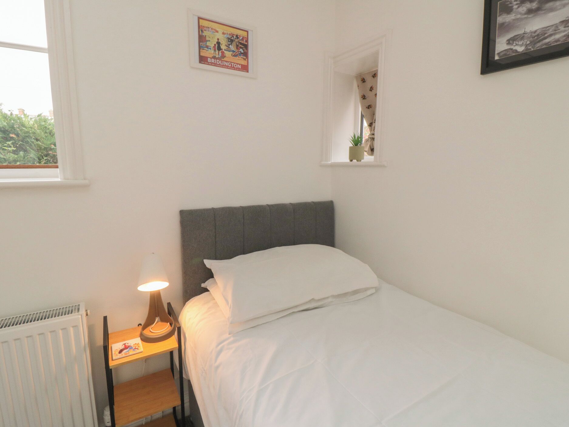 Une chambre avec un lit simple, une table de chevet avec une lampe, un radiateur, une fenêtre avec un rideau et un tableau encadré au mur.