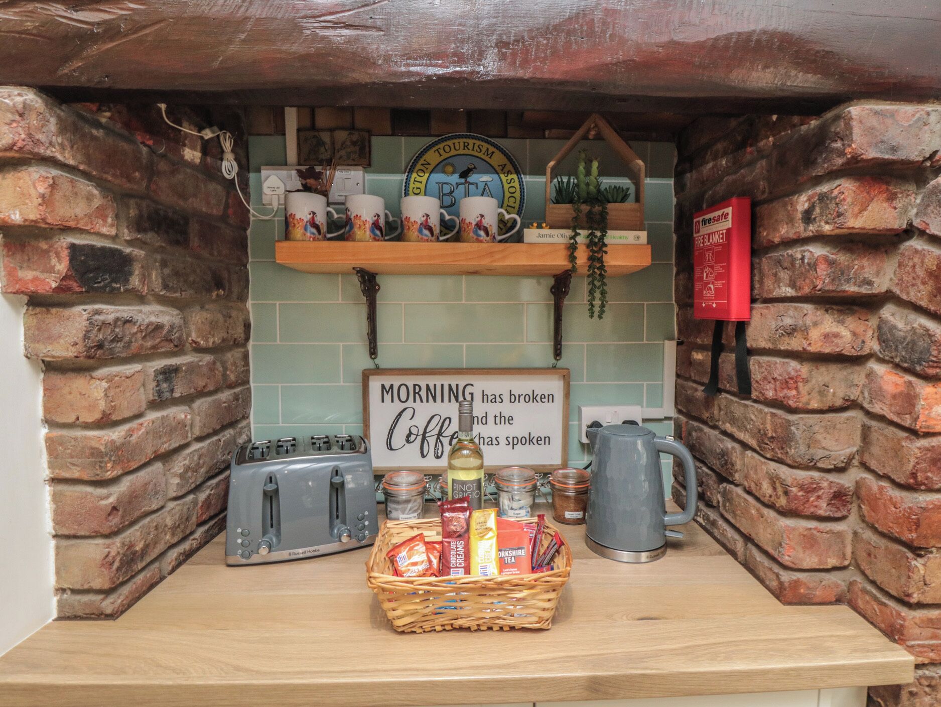 A kitchen counter with a sign, a toaster, a kettle, and a basket of tea and snacks.