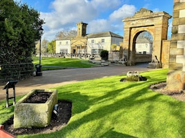 Un edificio histórico con un arco prominente, una torre de reloj y un césped cuidado con jardineras de piedra.
