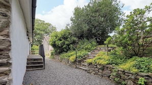 Una escalera de piedra que conduce a una puerta de madera, rodeada de abundante vegetación y un sendero de grava.