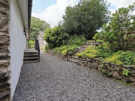 Una escalera de piedra que conduce a una puerta de madera, rodeada de abundante vegetación y un sendero de grava.