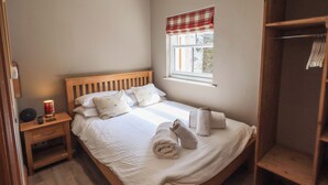 Una cama bien hecha con almohadas, una mesita de noche con lámpara, una ventana con cortinilla escocesa y un armario de madera.