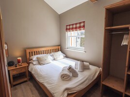 Una cama bien hecha con almohadas, una mesita de noche con lámpara, una ventana con cortinilla escocesa y un armario de madera.