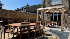 A patio with a wooden table and chairs, a pergola, and a stone building in the background. - The Oakroyd (Carnforth)