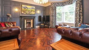 A living room with a fireplace, leather sofas, a chandelier, and a large window with curtains. - The Oakroyd (Carnforth)