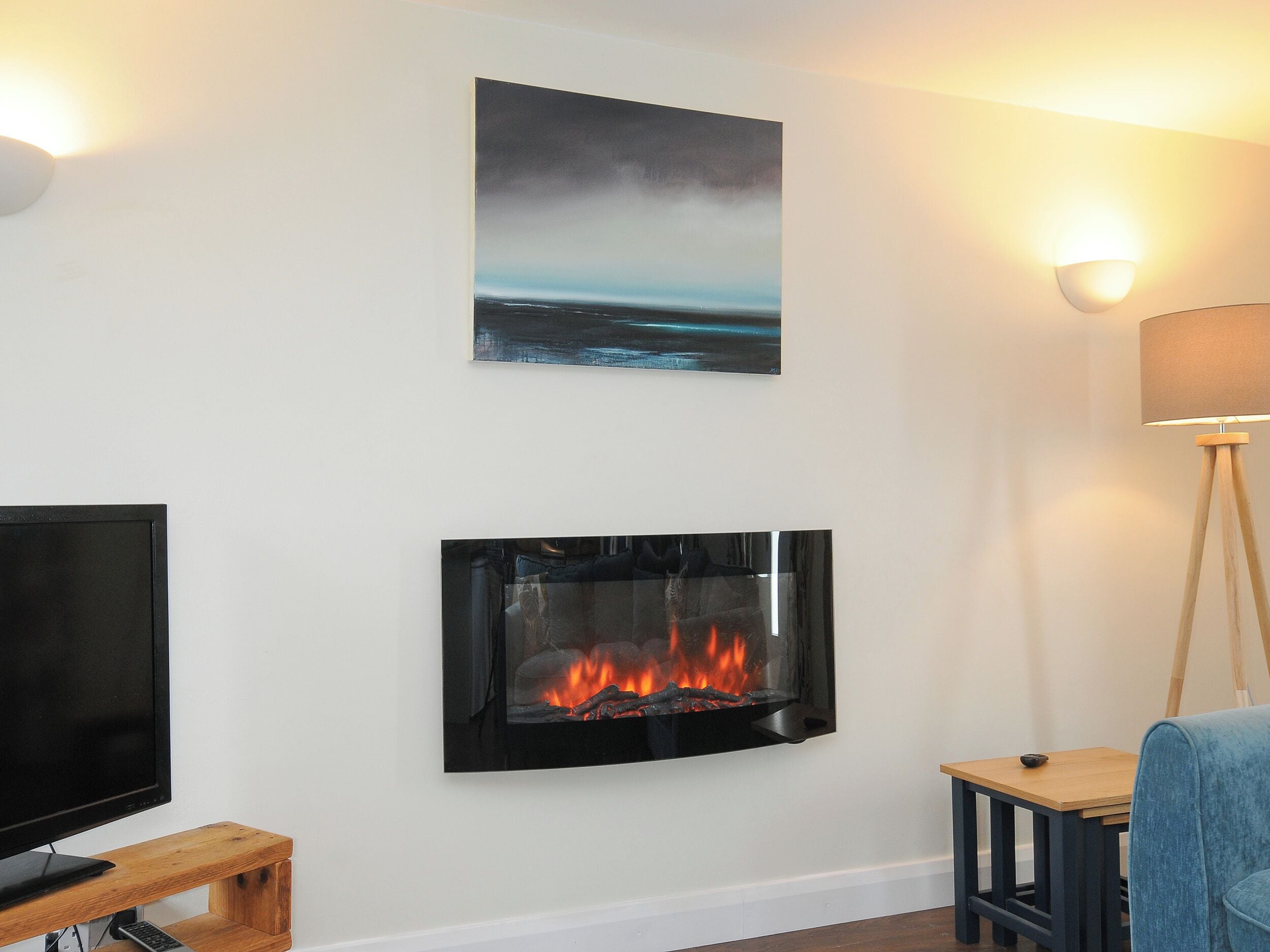 A living room with a flat-screen TV, a fireplace with visible flames, a painting of a seascape, and a floor lamp.