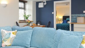Un saló modern amb un sofà blau, una taula de dinar amb caires i una cuina petita.