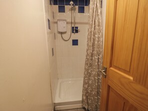 Een klein, afgesloten douchegedeelte met een witte betegelde wand, een handdouche en een douchegordijn met een bladmotief.